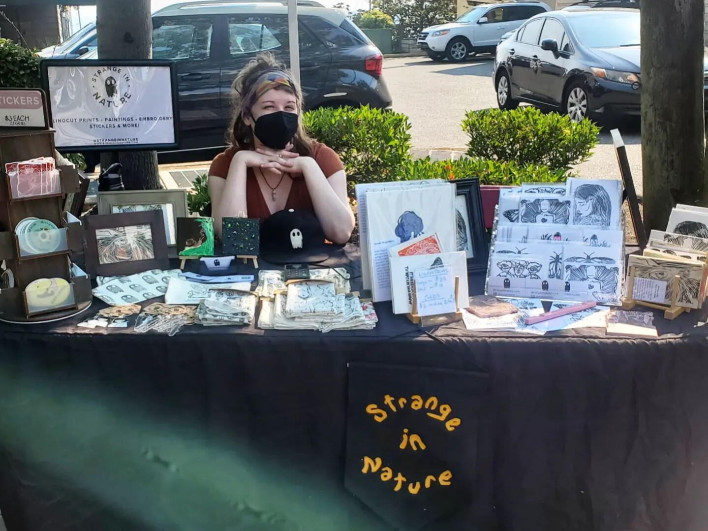 This is me at a market! A black table with the logo Strange in Nature in front of it. Displayed on the table are several hand-printed items made by Krista O'Halpin, the owner of Strange in Nature. She is seated happily behind the table, but is wearing a black medical